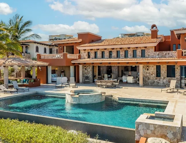 Luxurious villa with a stone facade overlooking a rectangular pool and jacuzzi. Palm trees and lounge chairs add a tropical, relaxing vibe.
