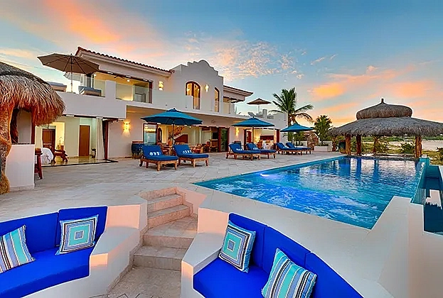 Modern luxury villa at sunset with a large pool, blue lounge chairs, and tropical umbrellas. Warm, inviting atmosphere with a clear sky backdrop.