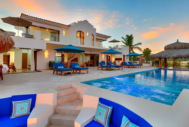 Modern luxury villa at sunset with a large pool, blue lounge chairs, and tropical umbrellas. Warm, inviting atmosphere with a clear sky backdrop.