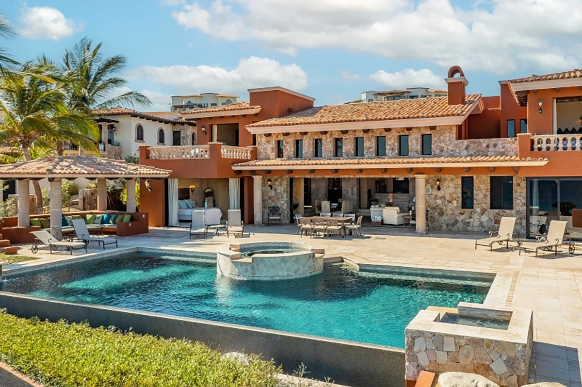 Luxurious villa with a stone facade overlooking a rectangular pool and jacuzzi. Palm trees and lounge chairs add a tropical, relaxing vibe.