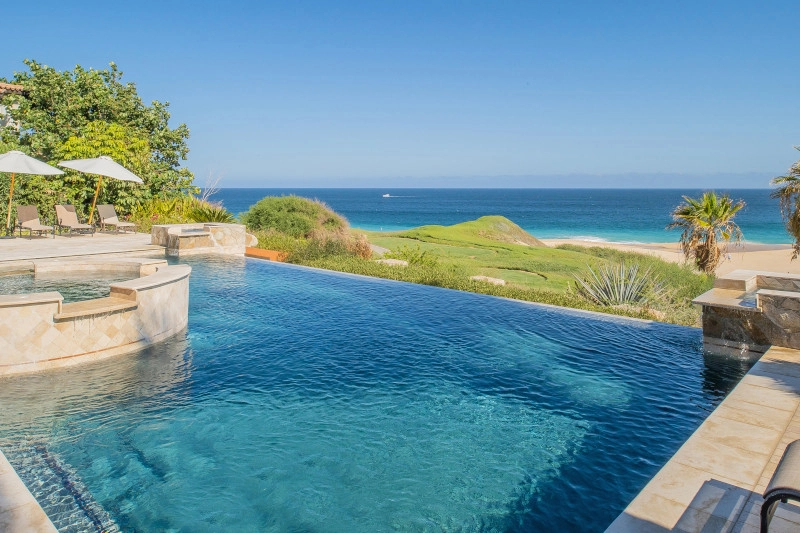 Infinity pool with a stone jacuzzi overlooking a green lawn, beach, and ocean under clear blue skies. Two lounge chairs with umbrellas on the left.
