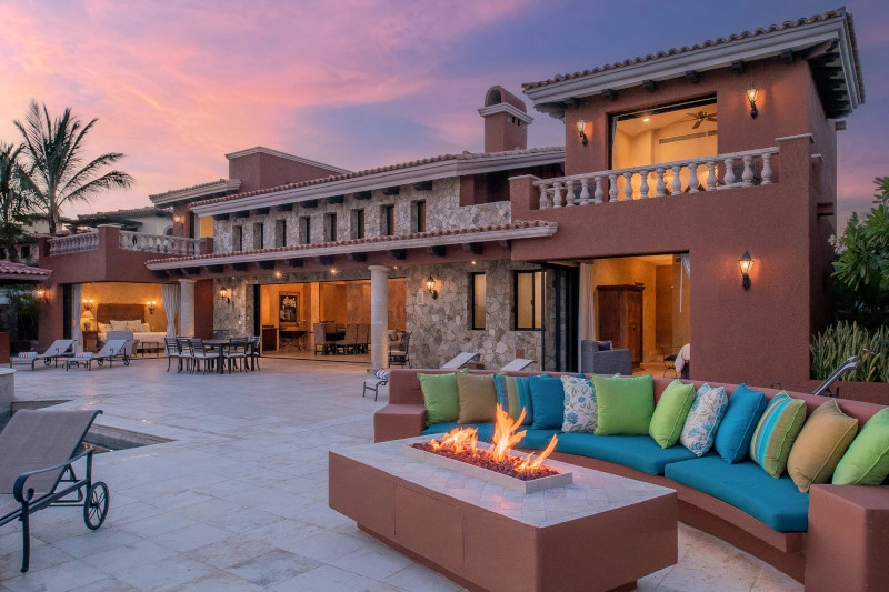 Spacious luxury villa at sunset with warm stone facade, palm trees, and an inviting patio. A lively fire pit is surrounded by vibrant cushions.